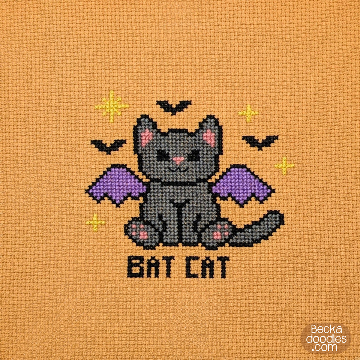 A cross-stitch design on orange Aida cloth showing a gray bat cat with purple wings, mini black bats, yellow stars, and the words “BAT CAT” stitched below.