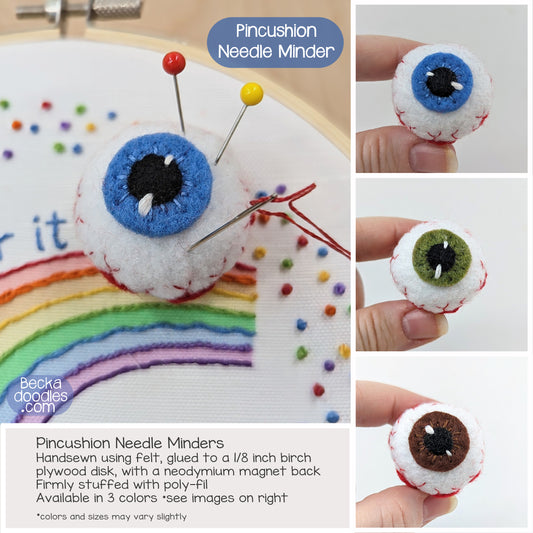 Pincushion needle minders with different colored eyes held by hands and used on a rainbow-themed embroidery project.