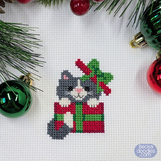 Cross-stitched cat inside a gift box with Christmas decorations on a white fabric background