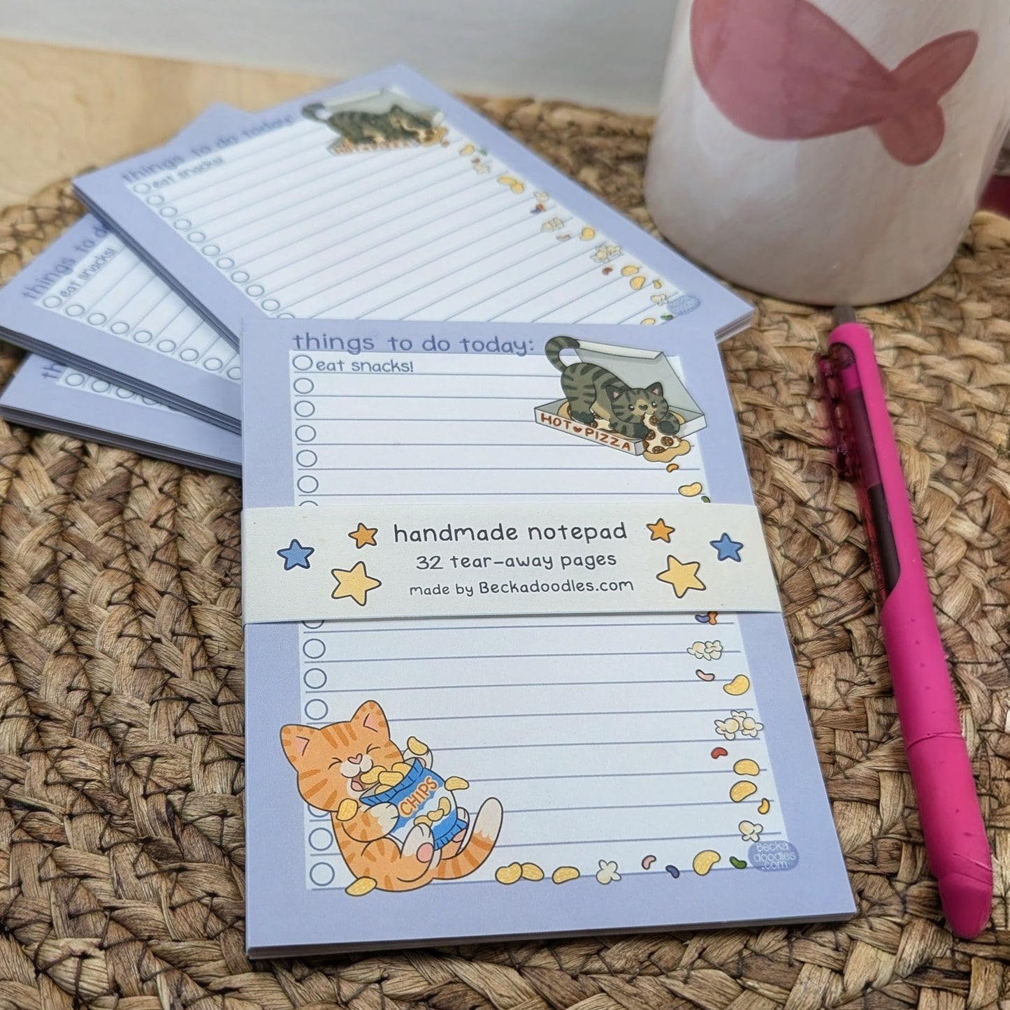 Handmade notepad with cat illustrations on a woven surface with a pink pen and mug.