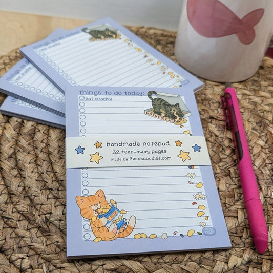 Handmade notepad with cat illustrations on a woven surface with a pink pen and mug.