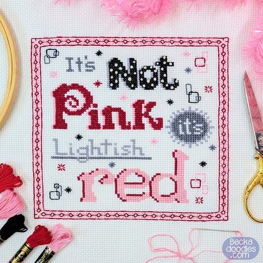 A square cross-stitch sampler with the pun 'It's Not Pink Its Lightish Red' stitched on white Aida fabric, surrounded by embroidery tools and pink and red thread.