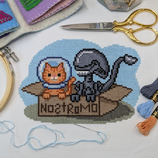 Cross-stitched design of a cat and alien in a space helmet with 'Nostromo' text, surrounded by embroidery tools.