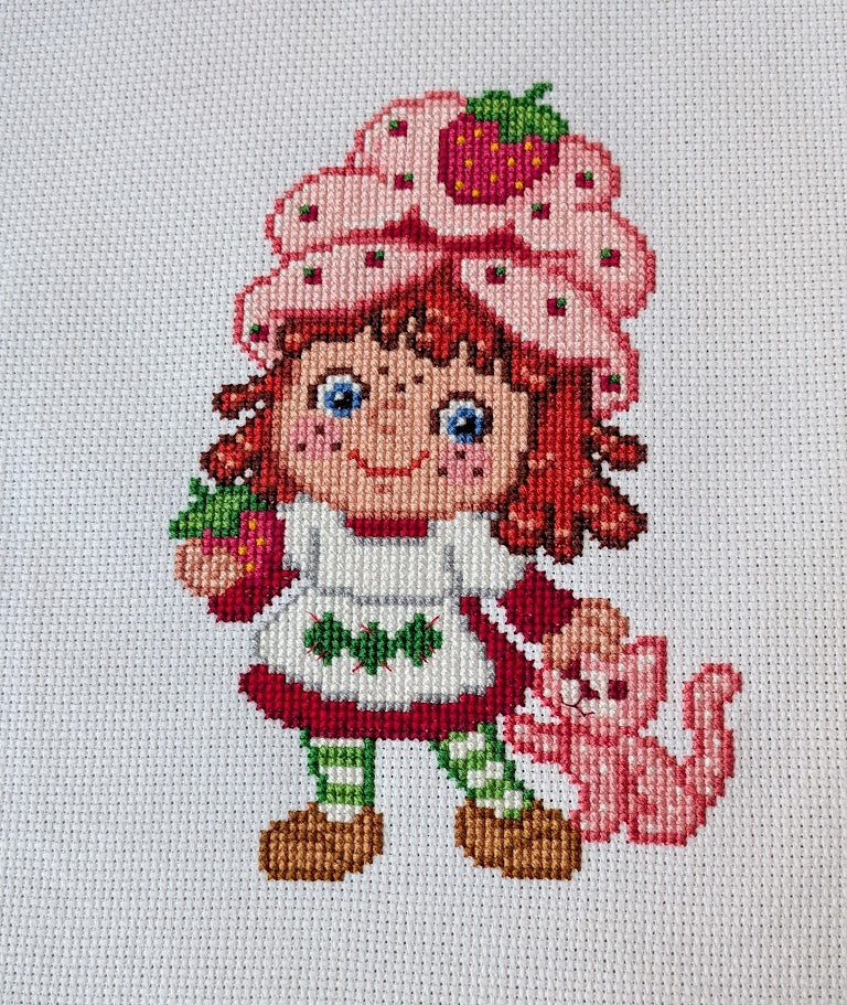 Cross-stitched design of a girl with red hair and a strawberry-themed hat, holding a pink cat.