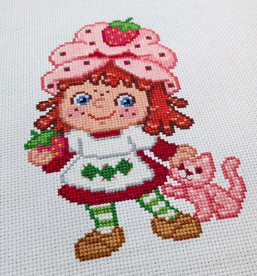 Cross-stitched character with a strawberry hat and pink cat on a white background