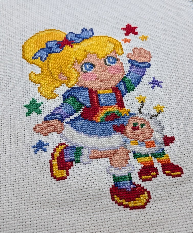 Cross-stitched design of a colorful character with stars and rainbow elements on a white background
