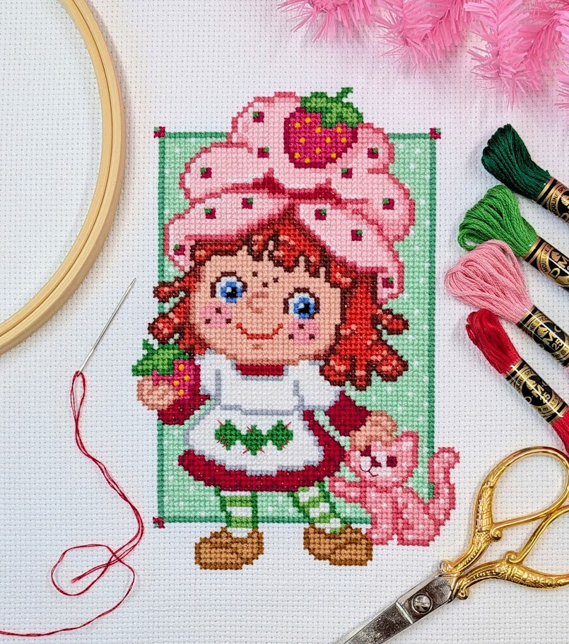 Cross-stitched design of a character with a strawberry hat, surrounded by embroidery threads and tools.