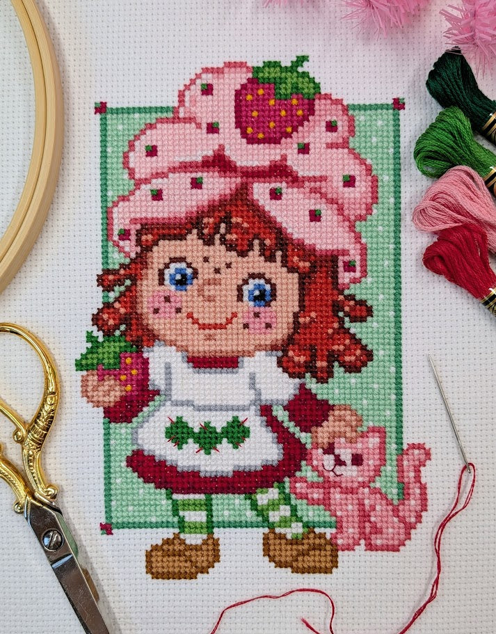 Cross-stitched design of a character with a pink hat and strawberry, surrounded by embroidery tools.