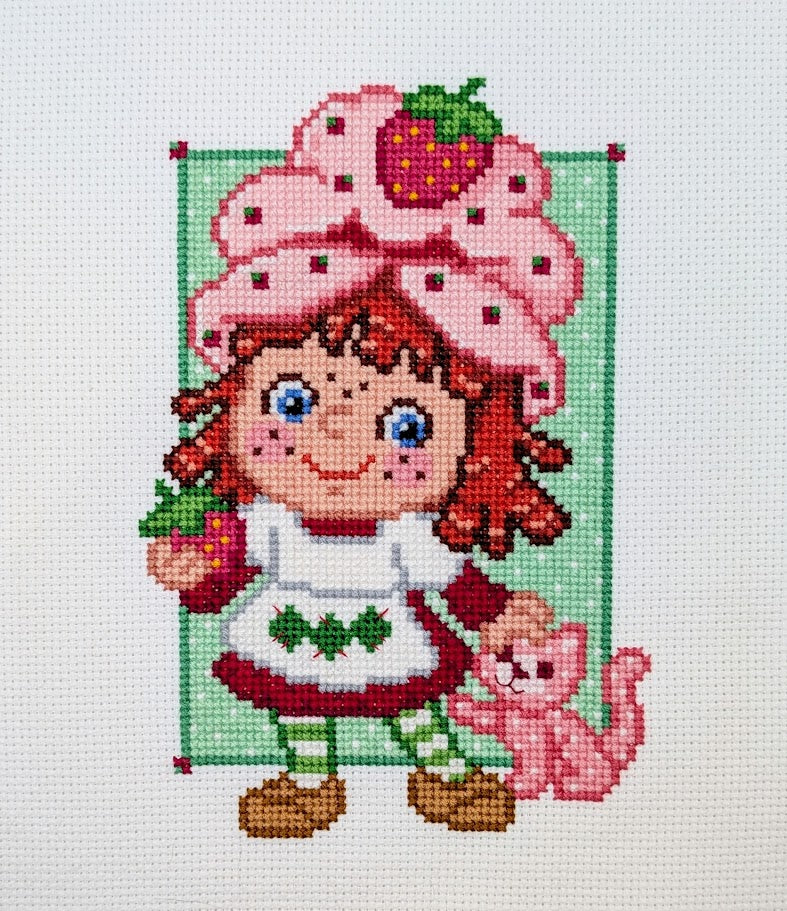 Cross-stitched design of a character with a strawberry-themed outfit and pink hat, holding a strawberry and a cat.