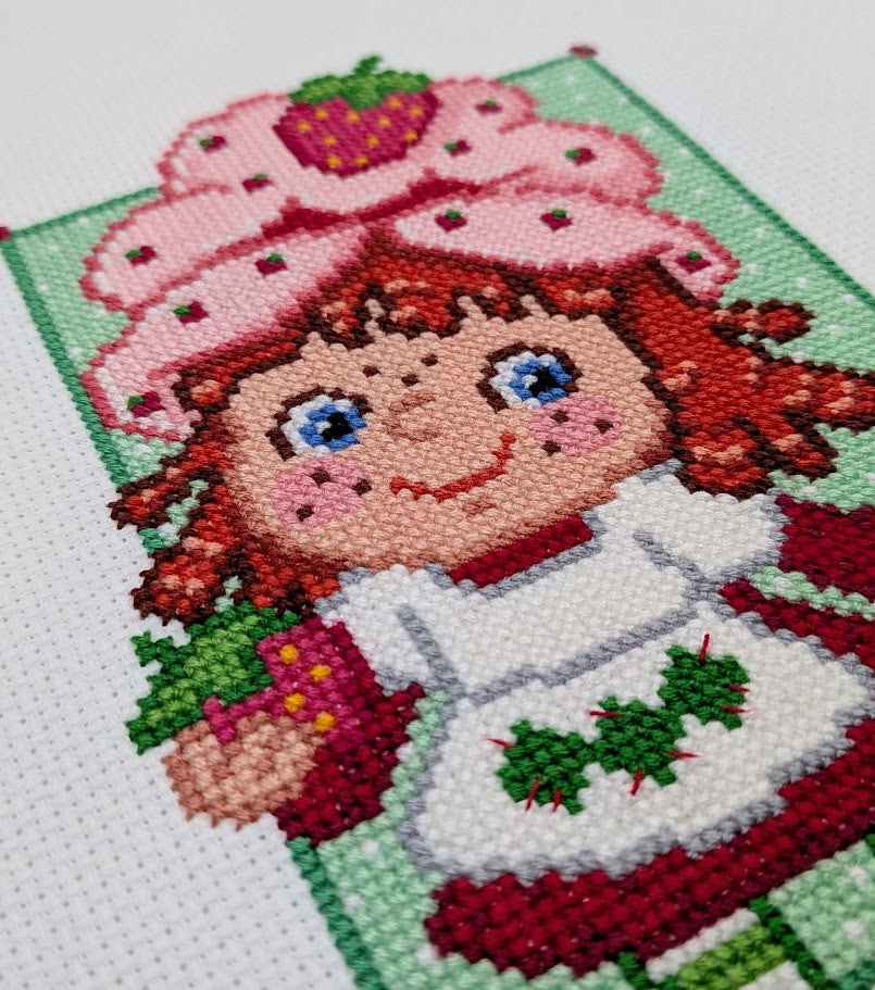 Cross-stitched design of a character with a strawberry hat and red hair on a white background