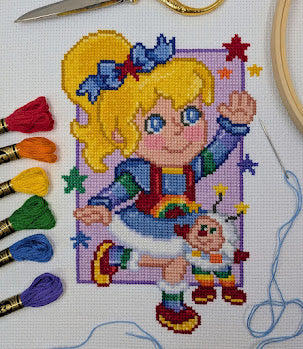 Cross-stitch pattern of a girl with rainbow-colored outfit and accessories, surrounded by embroidery threads and tools.