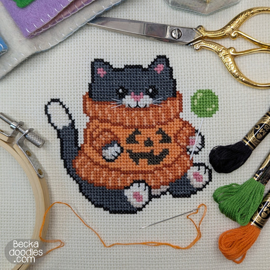 A completed cross-stitch design of a cute black cat wearing an orange pumpkin sweater, surrounded by scissors, embroidery floss, a needle, a hoop on a white Adia fabric. 