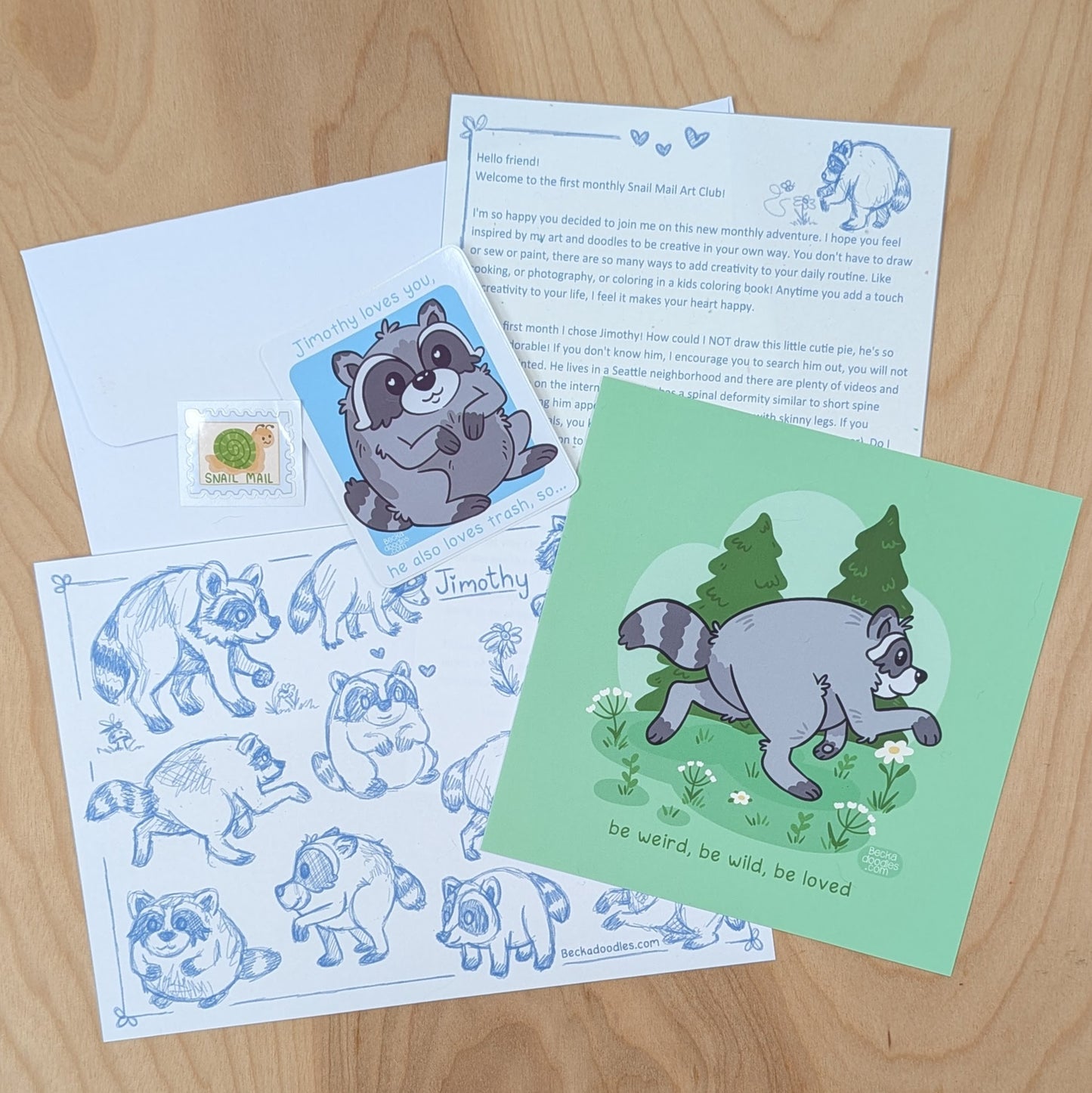 A set of raccoon-themed stationery items arranged on a light wood surface, including a green print, a blue illustrated sheet, an envelope, a printed letter, and a small sticker.