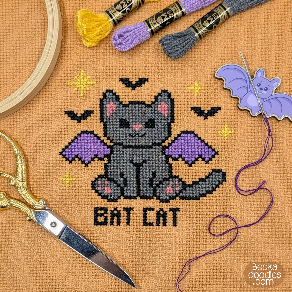 A cross-stitch design of a gray bat cat with purple wings and the words 'BAT CAT' stitched below, stitched on orange Aida cloth with embroidery hoops, scissors, thread, and a bat-shaped needle holder.