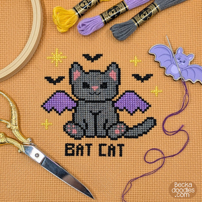 A cross-stitch design of a gray bat cat with purple wings and the words 'BAT CAT' stitched below, stitched on orange Aida cloth with embroidery hoops, scissors, thread, and a bat-shaped needle holder.