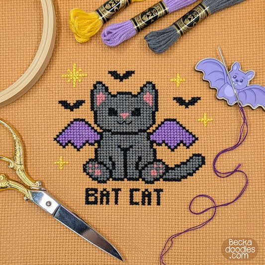 A cross-stitch design of a gray bat cat with purple wings and the words 'BAT CAT' stitched below, stitched on orange Aida cloth with embroidery hoops, scissors, thread, and a bat-shaped needle holder.