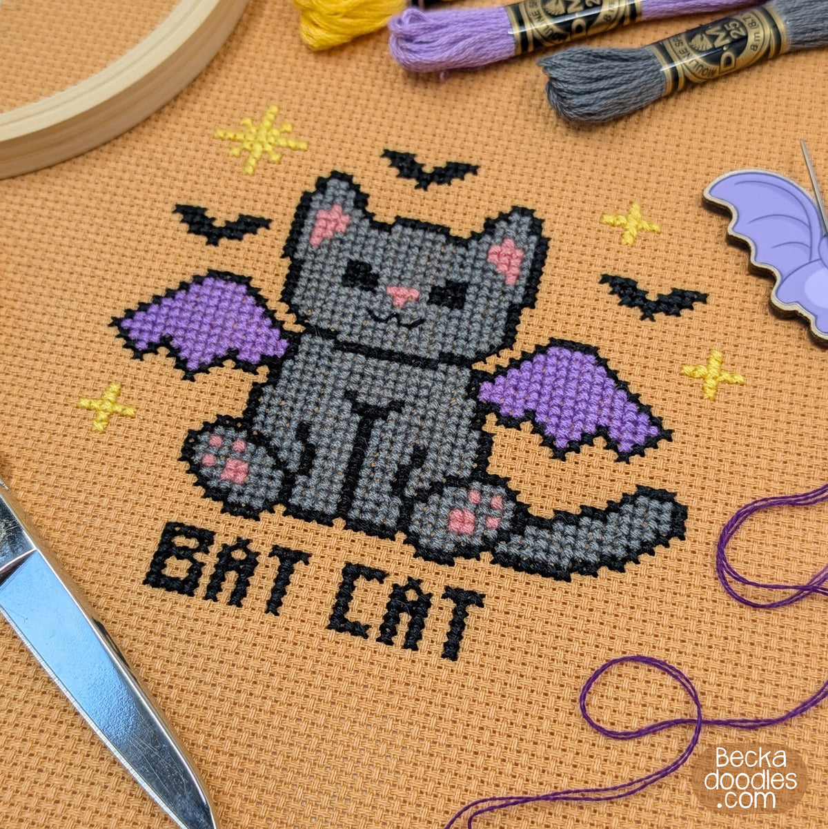 A close-up of a cross-stitch design on orange Aida cloth, showing a gray cat with purple wings and the words 'BAT CAT' stitched below. Surrounding by embroidery floss, a wooden hoop, and scissors.