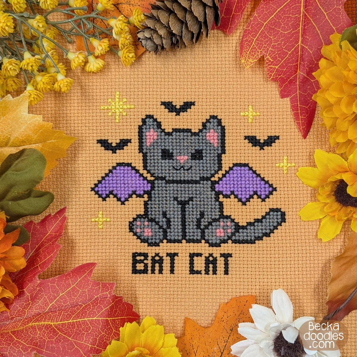A cross-stitch design of a gray bat cat with purple wings and the words 'BAT CAT' stitched below, surrounded by autumn leaves, pinecones, and flowers on a orange Aida cloth.