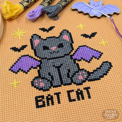 A close-up of a cross-stitch design featuring a gray bat cat with purple wings, pink inner ears and paws, and the words “BAT CAT” stitched below. Stitched on orange Aida cloth with embroidery floss, a bat-shaped needle minder, and needle and thread.