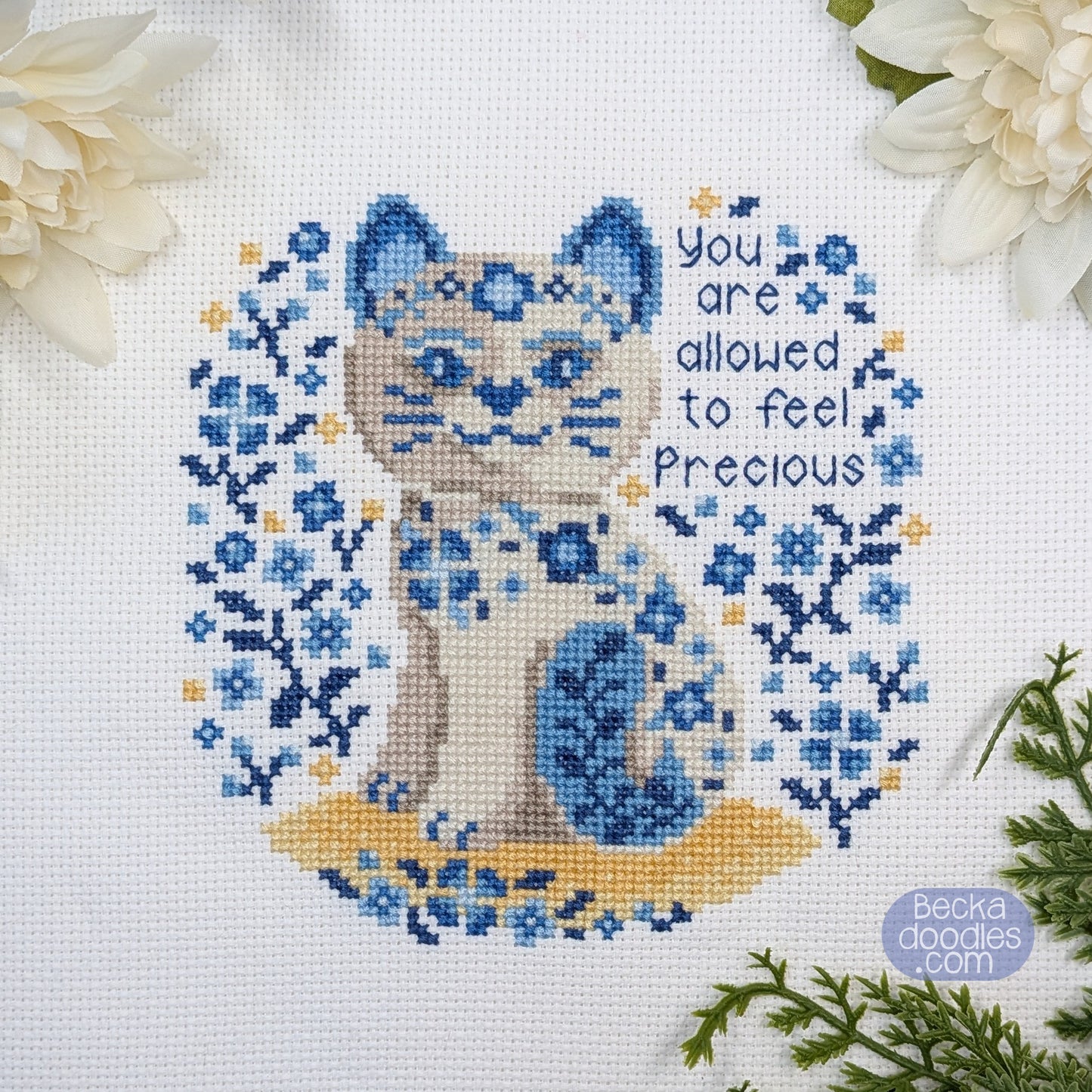 Cross-stitched porcelain cat design with floral patterns and text 'You are allowed to feel precious' on a white Adia cloth.