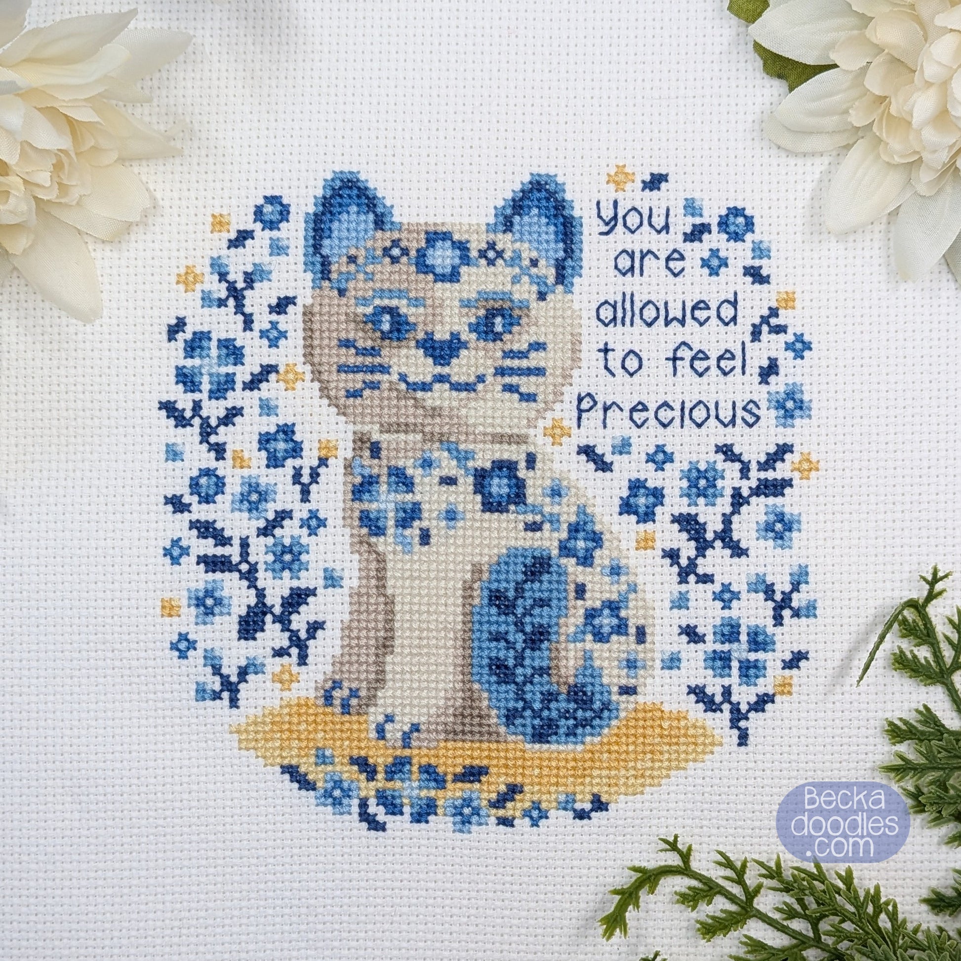 Cross-stitched porcelain cat design with floral patterns and text 'You are allowed to feel precious' on a white Adia cloth.