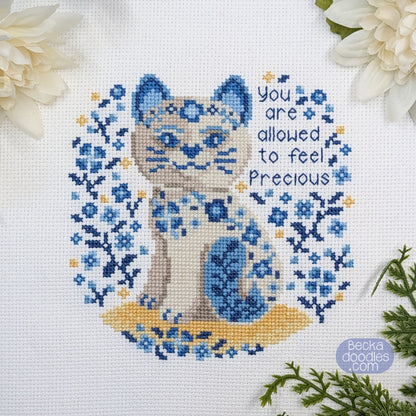 Cross-stitched porcelain cat design with floral patterns and text 'You are allowed to feel precious' on a white Adia cloth.