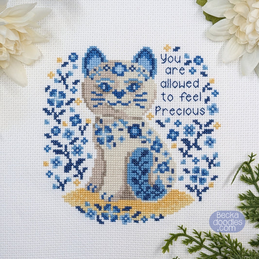 Cross-stitched porcelain cat design with floral patterns and text 'You are allowed to feel precious' on a white Adia cloth.