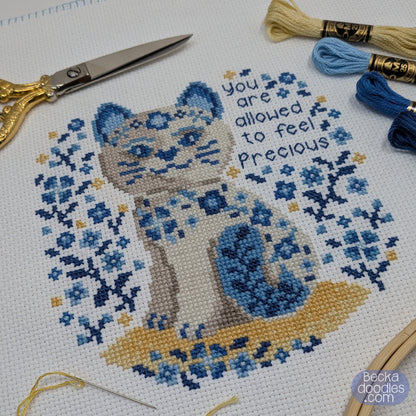 Cross-stitched design of a porcelain cat with floral patterns and text 'you are allowed to feel Precious' on fabric, with embroidery tools around.