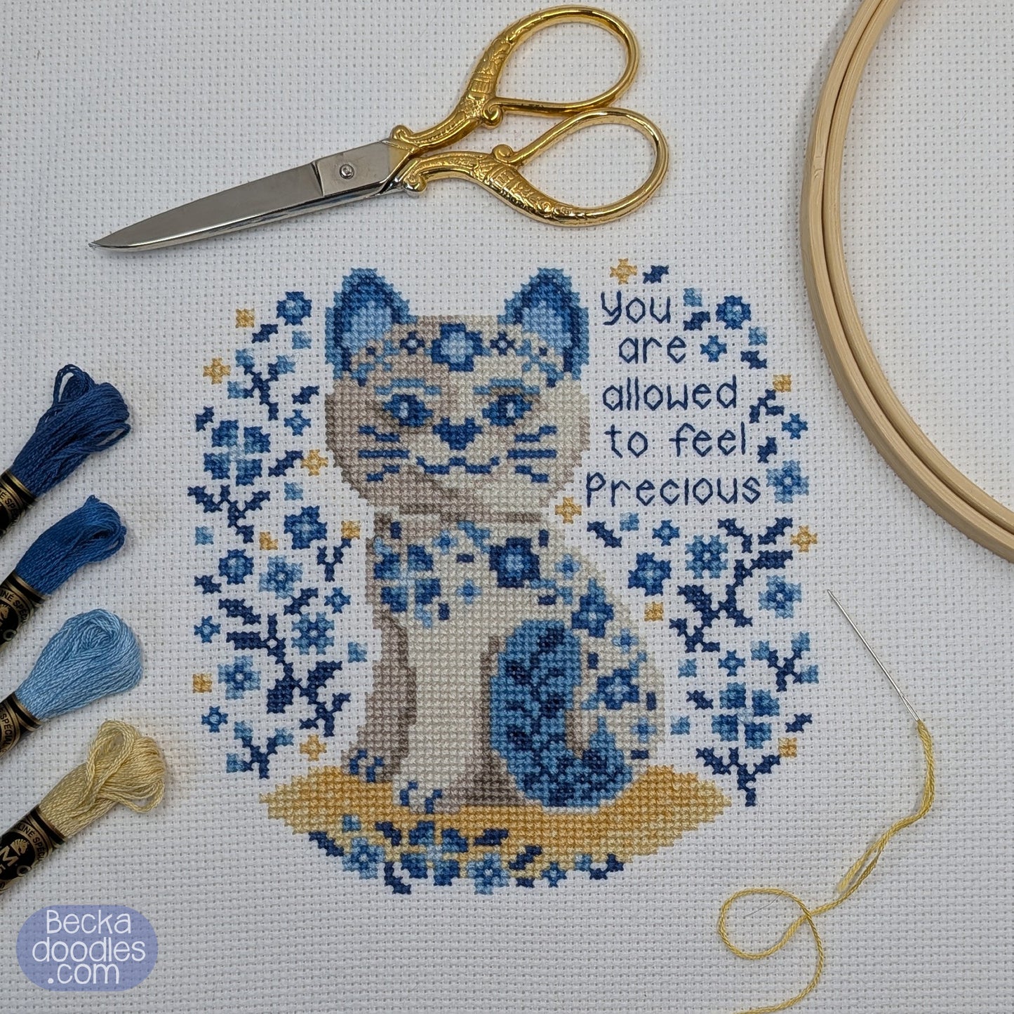 Cross-stitched porcelain cat design with decorative text, embroidery tools, and hoop on a white fabric background.