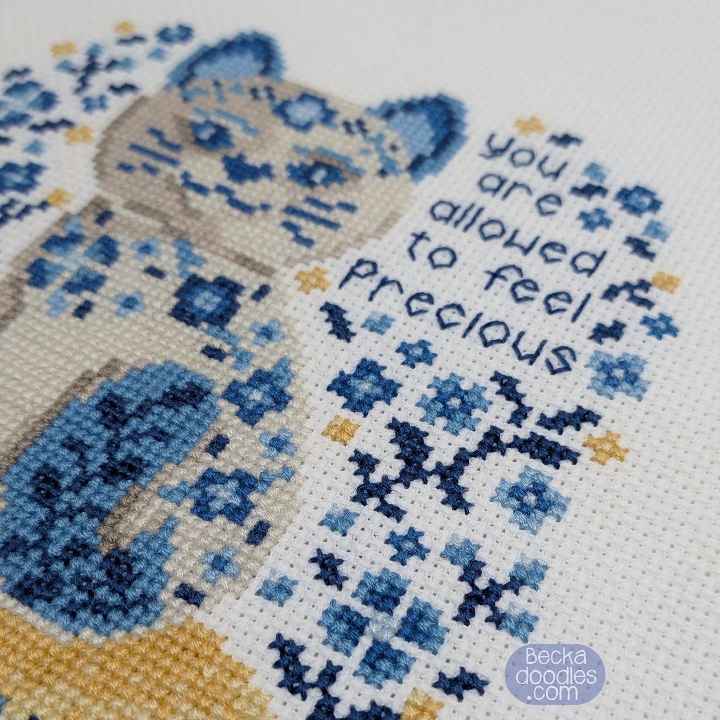 Cross-stitched design of a cat with flowers and text 'you are allowed to feel precious' on a white Adia fabric.