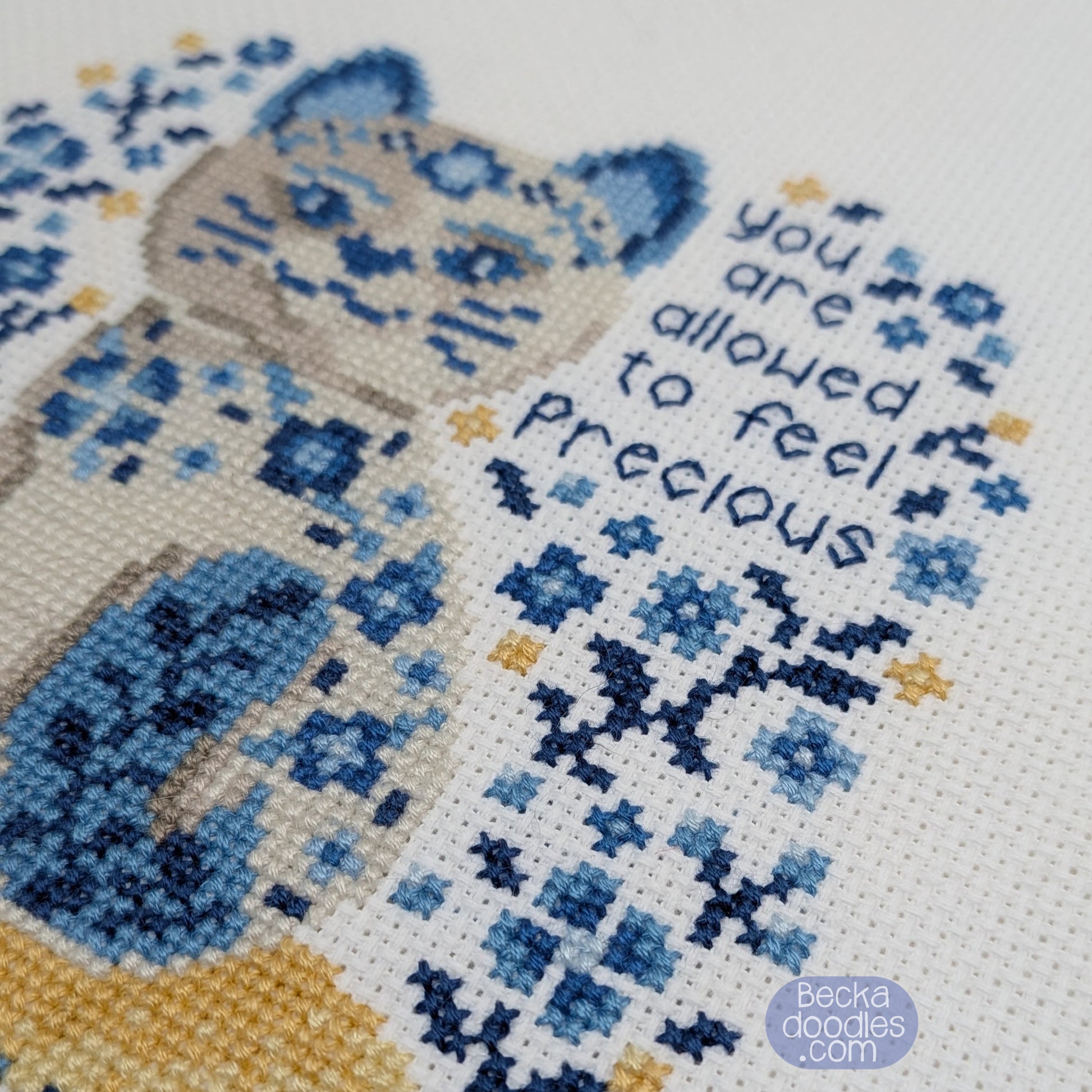 Cross-stitched design of a cat with flowers and text 'you are allowed to feel precious' on a white Adia fabric.