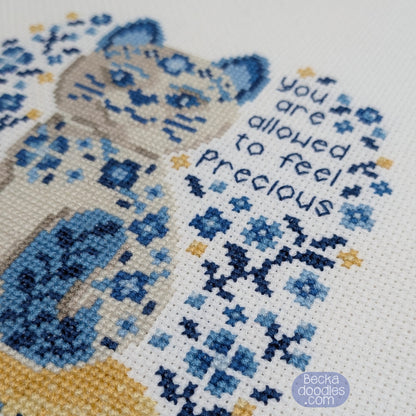 Cross-stitched design of a cat with flowers and text 'you are allowed to feel precious' on a white Adia fabric.