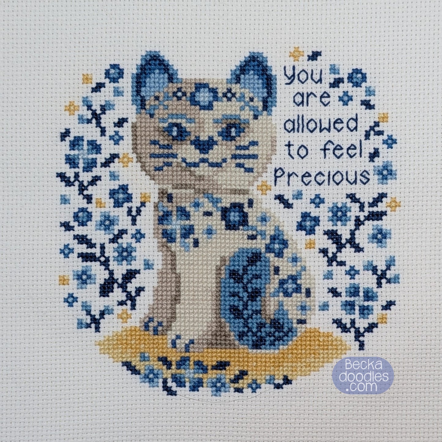 Cross-stitched design of a porcelain cat surrounded by flowers with text 'You are allowed to feel precious' on a white fabric.