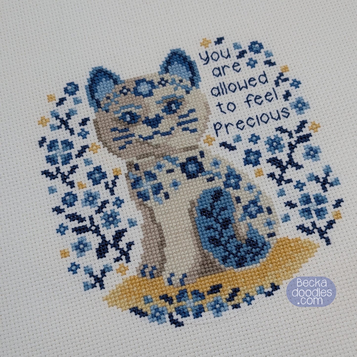 Cross-stitched design of a porcelain cat with floral patterns and text on white Aida fabric.