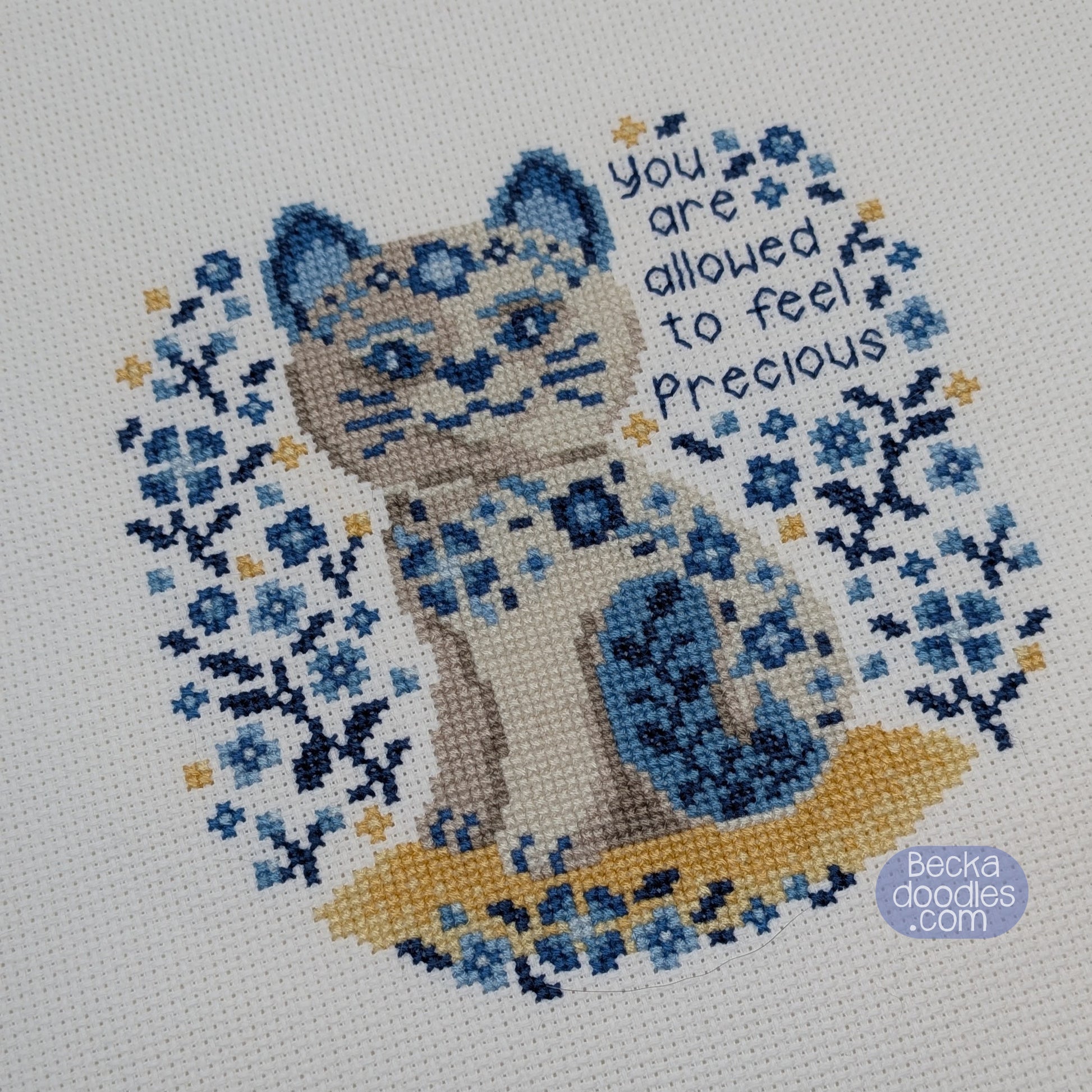 Cross-stitched design of a porcelain cat with floral patterns and text on white Aida fabric.