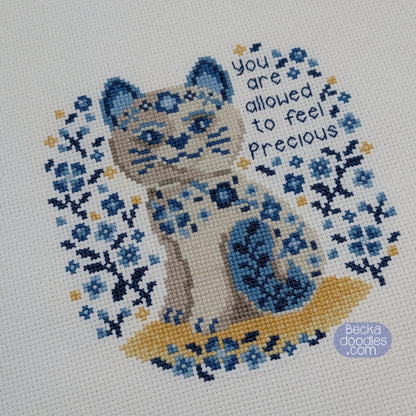 Cross-stitched design of a porcelain cat with floral patterns and text on white Aida fabric.