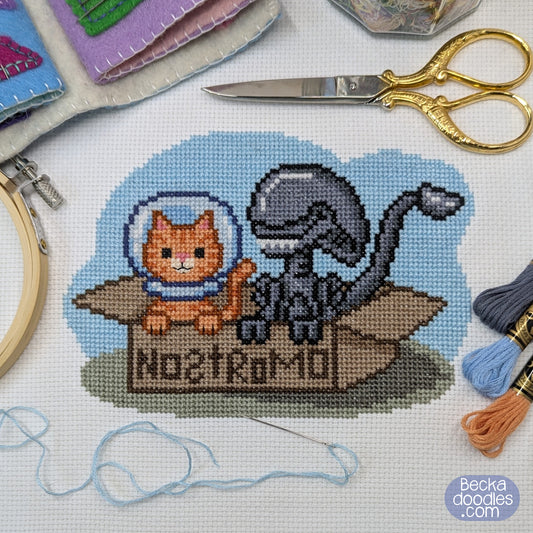 Cross-stitch design on white fabric showing an orange cat in a space helmet next to a gray alien sitting in a box labeled 'NOSTROMO'. Surrounded by scissors, embroidery hoop, thread, and other sewing supplies.