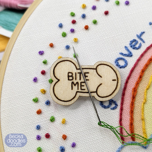 Embroidery hoop with colorful stitches and a wooden penis with 'BITE ME' text.