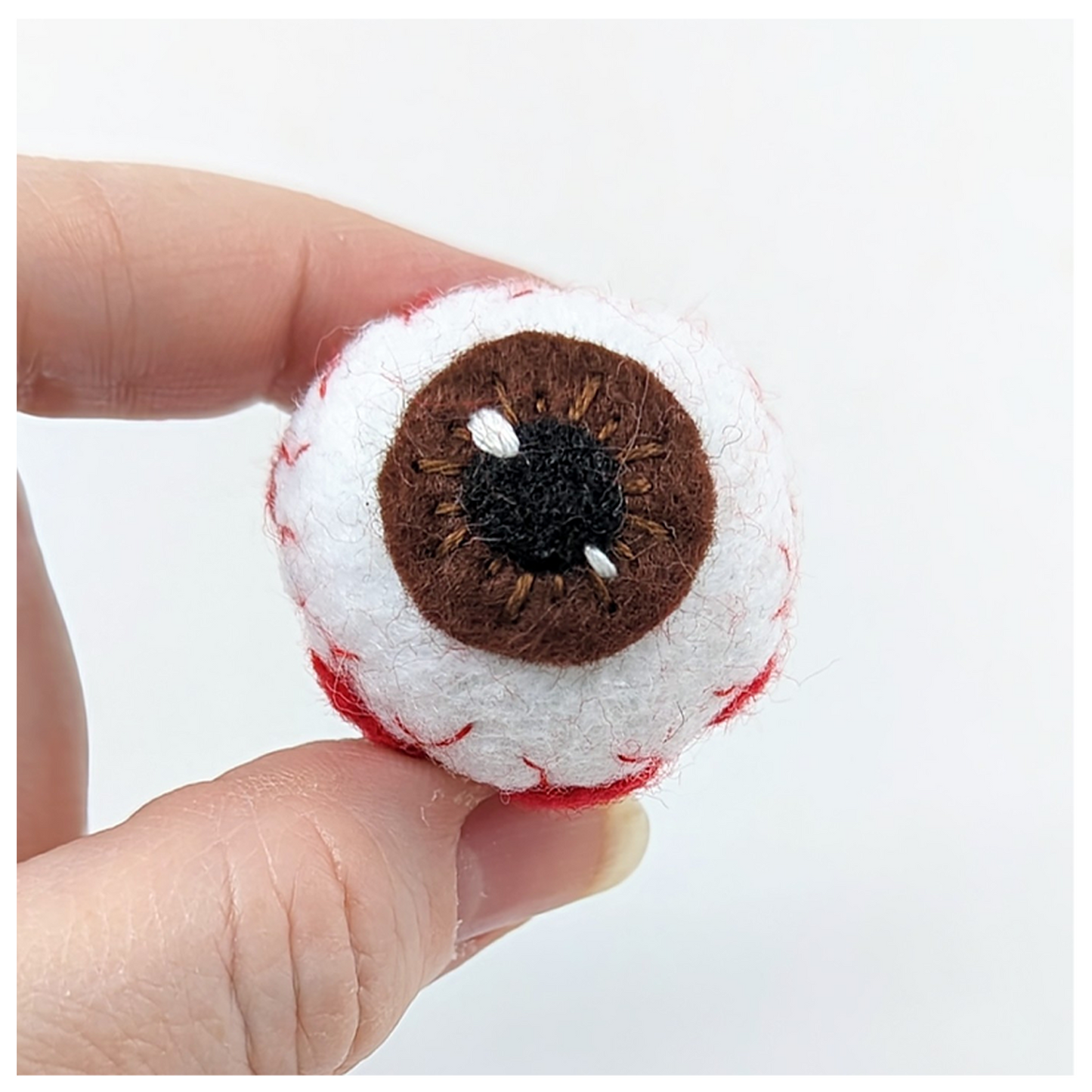 Felted eye with brown iris held between fingers against a white background
