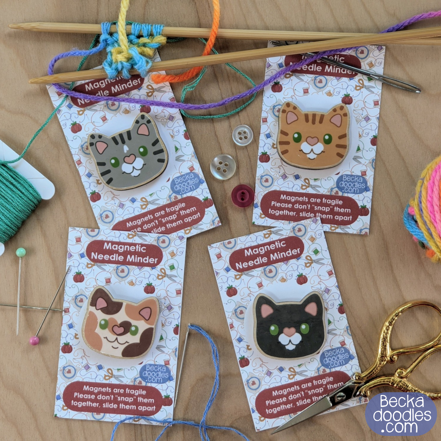 Four cat-themed magnetic needle minders on a wooden surface with knitting needles and yarn.
