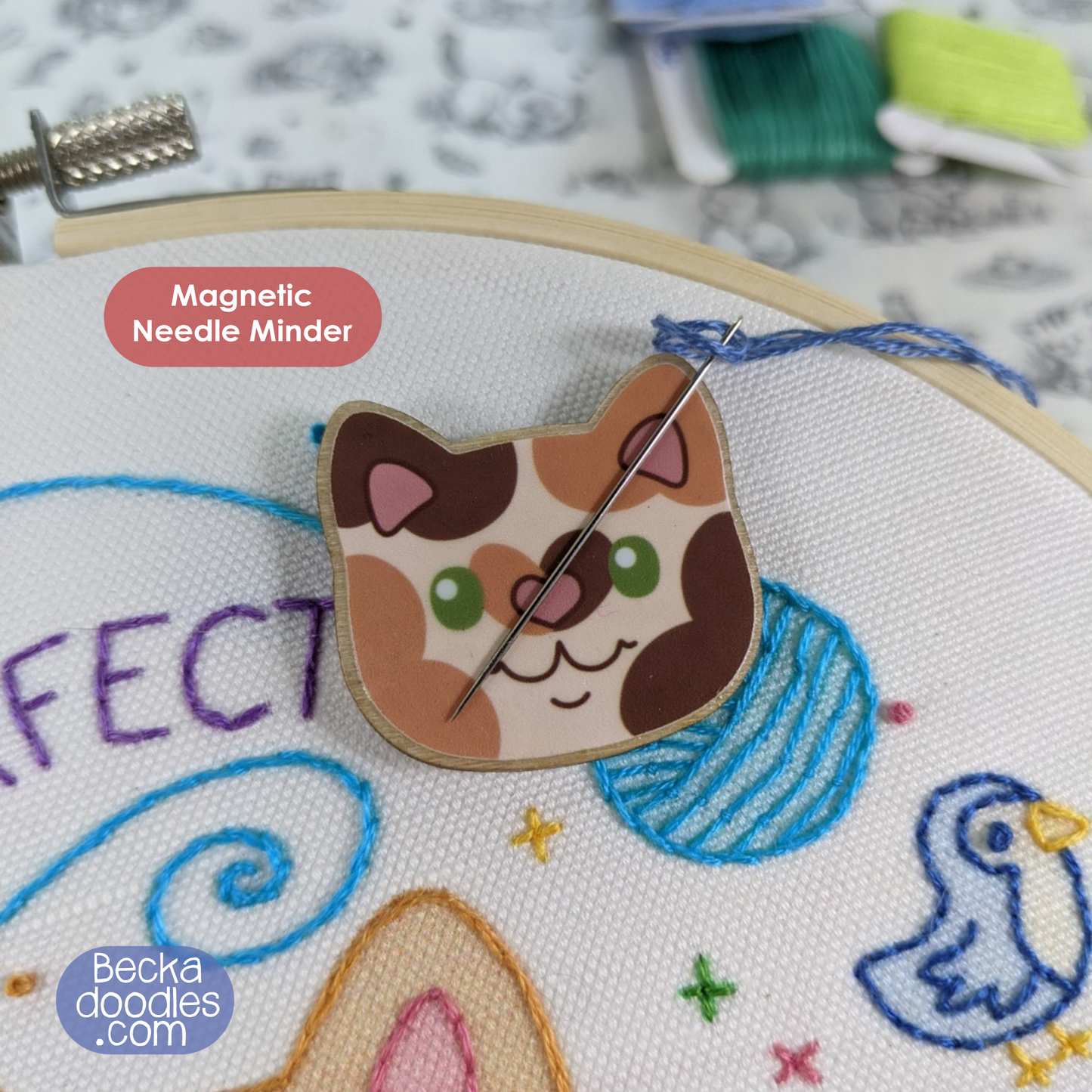 Cat-shaped magnetic needle minder on an embroidery hoop with colorful stitches.