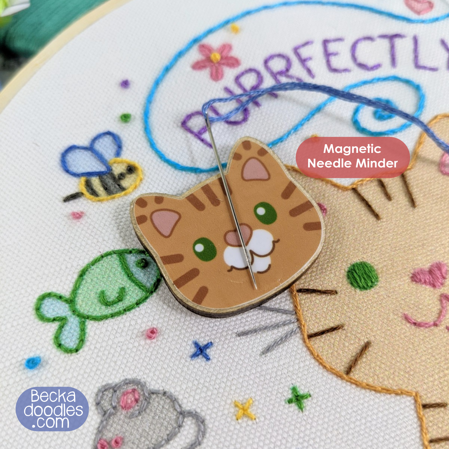 Cat-shaped magnetic needle minder on an embroidery hoop with colorful stitches and text.