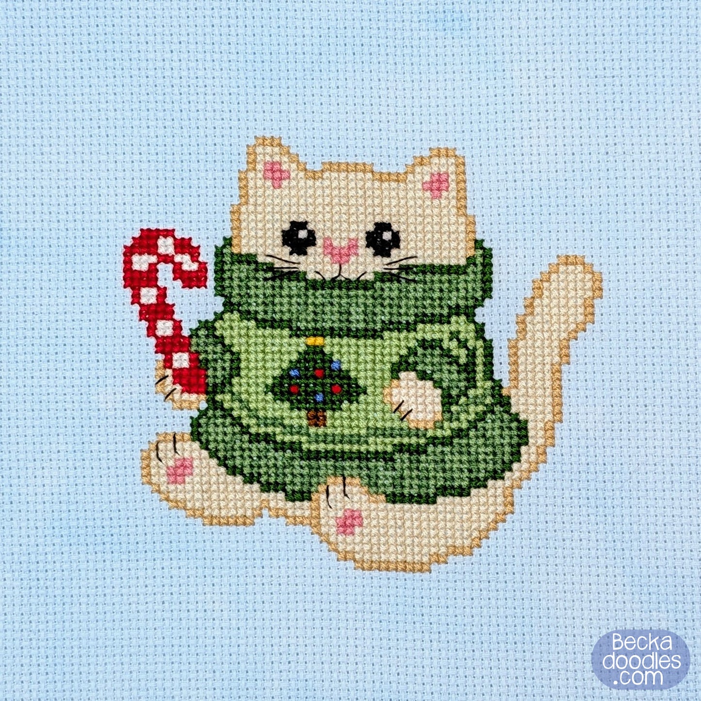 A cross-stitch design of a cream colored cat wearing a green sweater and holding a red-and-white candy cane, on light blue Adia fabric. 