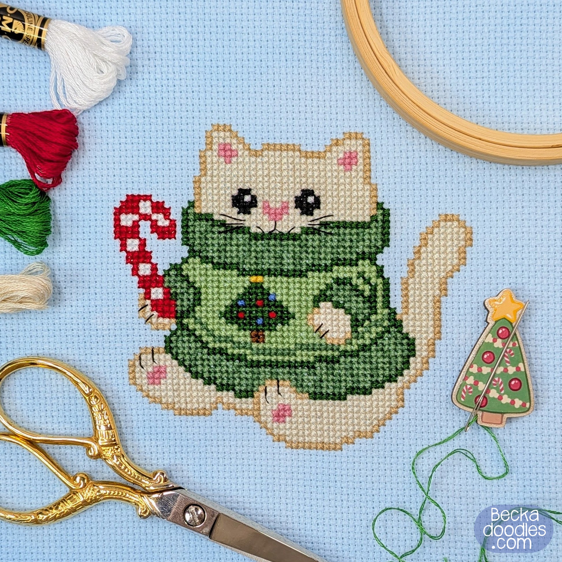 A completed cross-stitch design of a cream-colored cat wearing a green sweater with a small Christmas tree on the chest, holding a red-and-white candy cane. The piece is displayed on light blue Adia fabric with embroidery tools and thread around it, including scissors, thread, an embroidery hoop edge, and a small Christmas needle holder.