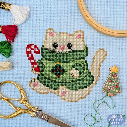 A completed cross-stitch design of a cream-colored cat wearing a green sweater with a small Christmas tree on the chest, holding a red-and-white candy cane. The piece is displayed on light blue Adia fabric with embroidery tools and thread around it, including scissors, thread, an embroidery hoop edge, and a small Christmas needle holder.