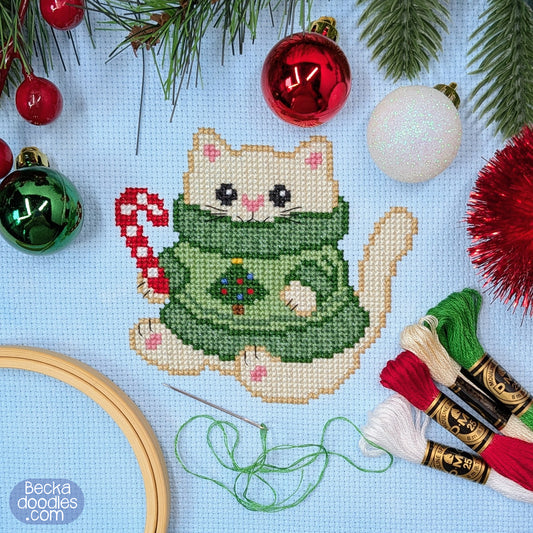 A festive cross-stitch design featuring a cute cream-colored cat wearing a green Christmas sweater, surrounded by embroidery floss, a wooden hoop, pine branches, and red and green ornaments on a light blue fabric. 