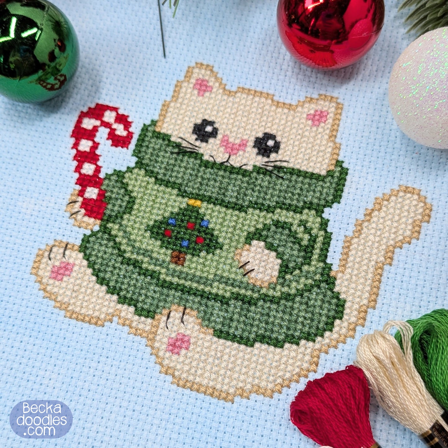 A completed cross-stitch design of a cute cream-colored cat wearing a green Christmas sweater and holding a red-and-white candy cane, displayed on a light blue fabric with festive ornaments and thread skeins around it. 