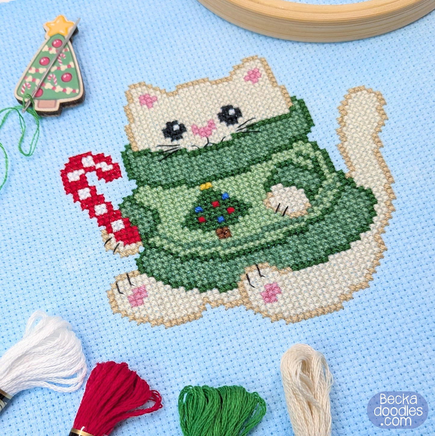 A Christmas-themed cross-stitch design of a cream colored cat wearing a green sweater with a small Christmas tree on it, holding a red-and-white candy cane. The finished design is stitched on light blue fabric with embroidery thread, a wooden hoop, and a decorative Christmas tree needle holder around the design.