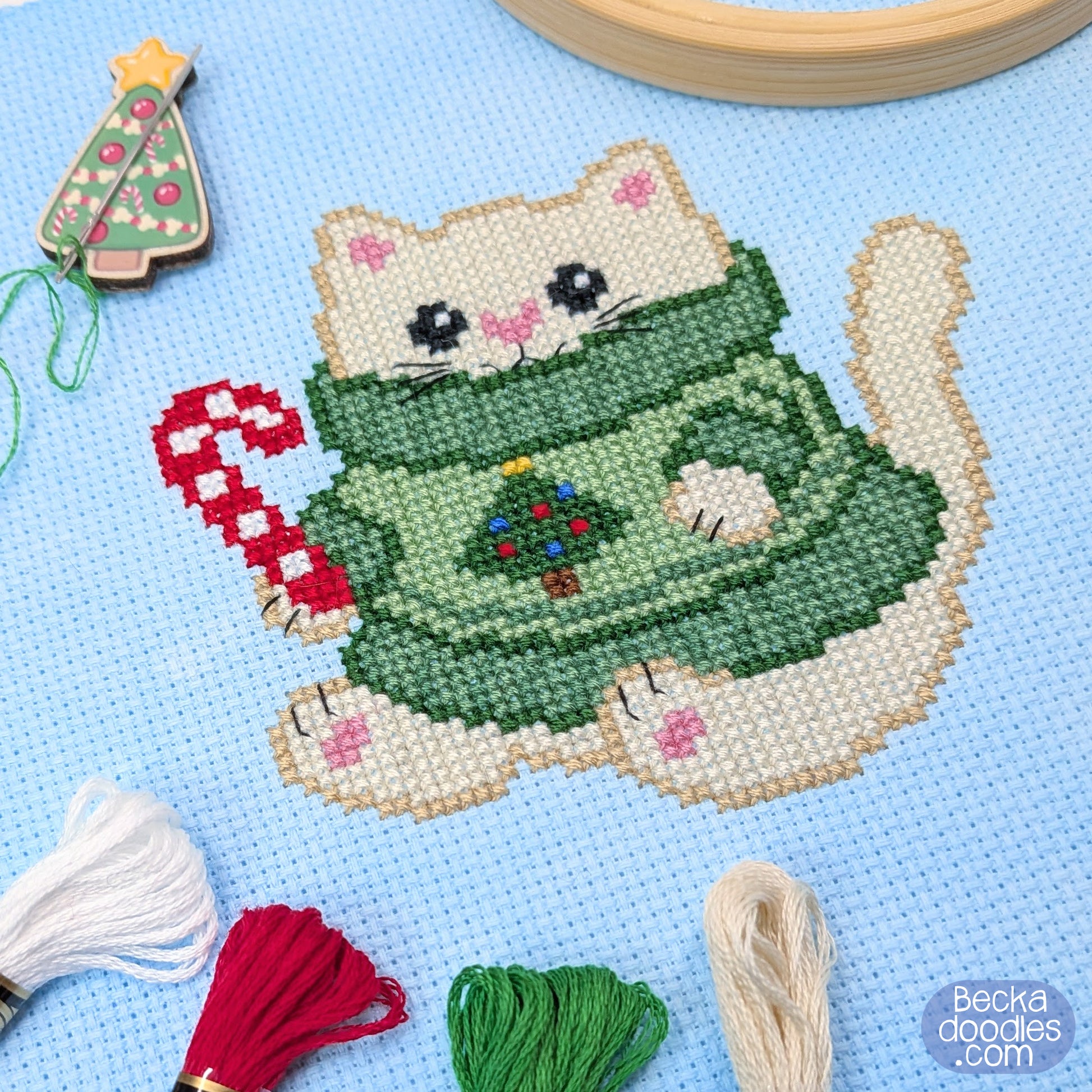 A Christmas-themed cross-stitch design of a cream colored cat wearing a green sweater with a small Christmas tree on it, holding a red-and-white candy cane. The finished design is stitched on light blue fabric with embroidery thread, a wooden hoop, and a decorative Christmas tree needle holder around the design.
