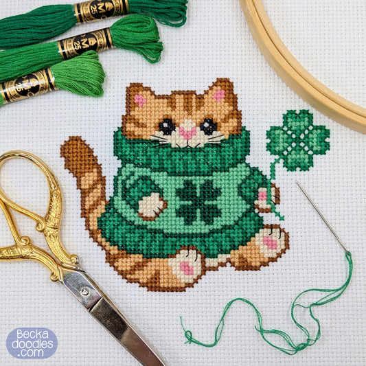 Cross-stitch pattern of a cat in a green sweater with a four-leaf clover, surrounded by embroidery tools on white fabric.